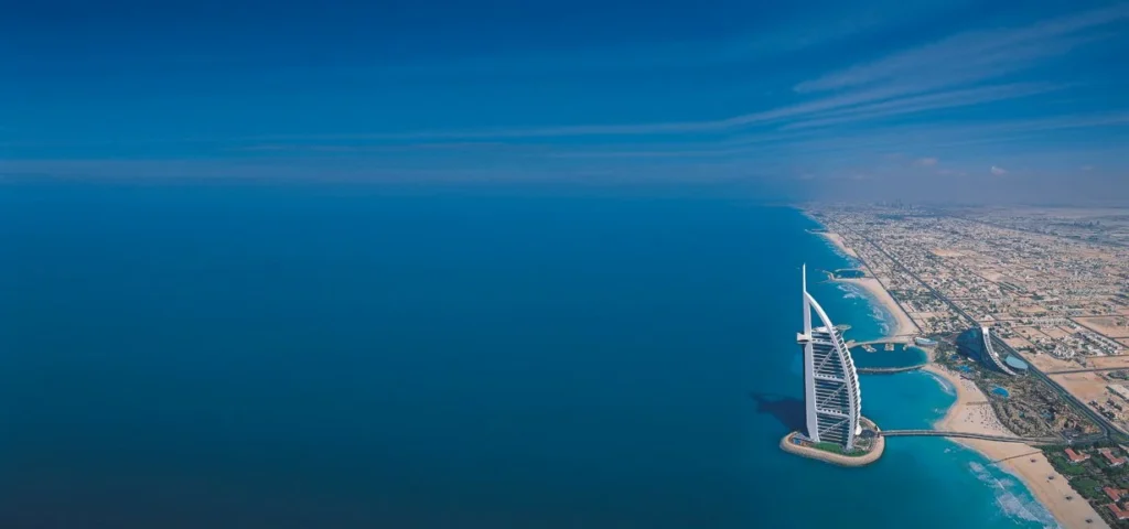 Aerial view of the Burj Al Arab hotel standing on its man-made island along Dubai’s coastline, surrounded by the vast blue waters of the Arabian Gulf.