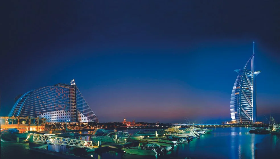 A night-time marina view featuring the Burj Al Arab illuminated against the Dubai skyline, with yachts docked in the foreground.