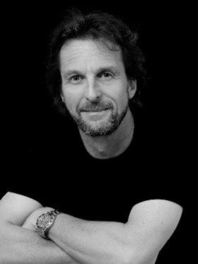 Portrait of an Tom Wright architect with medium-length hair and a trimmed beard, wearing a black T-shirt and wristwatch, posing confidently against a black background.