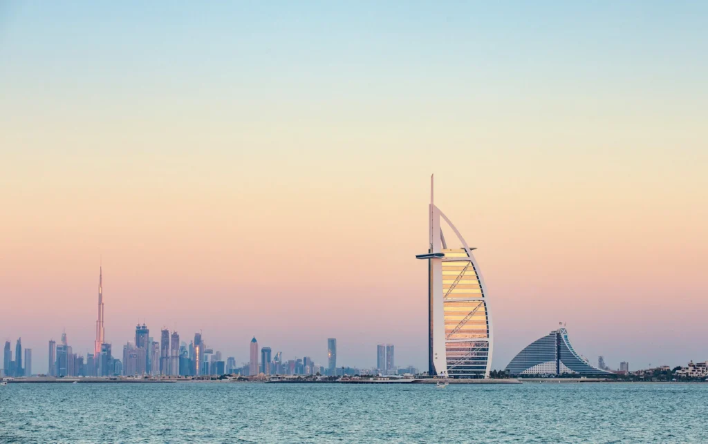 The Burj Al Arab seen from the sea during sunset, with the Dubai skyline— including the Burj Khalifa—visible in the background.