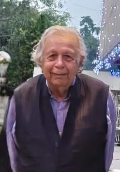 Senior man standing outdoors in evening lighting, dressed in a dark vest and shirt, looking at the camera with a gentle expression.