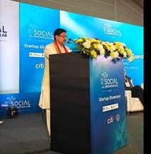 A speaker addressing the audience at a podium during a Social Innovation Lab event, with a blue event backdrop and floral arrangement on the podium.