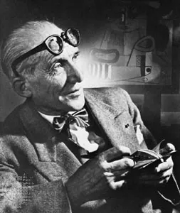 Black-and-white portrait of architect Le Corbusier wearing round glasses on his forehead and holding a small book.