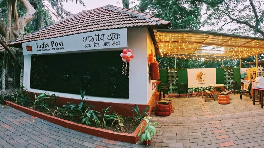 Exterior view of the Gen-Z themed Post Office Extension Counter at CMS College, featuring a landscaped entrance, India Post signage, and a semi-open seating zone with ambient lighting.