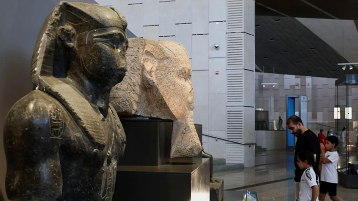 Granite statues of ancient Egyptian pharaohs displayed inside the Grand Egyptian Museum with visitors exploring the gallery.
