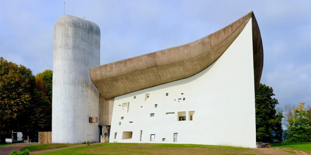 Exterior view of Notre-Dame du Haut chapel in Ronchamp, France, featuring its sculptural curving roof and white textured walls.