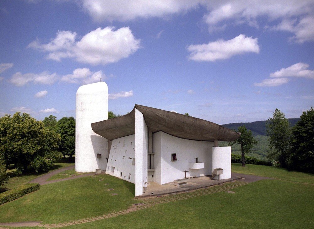 Wide-angle view of the Ronchamp chapel surrounded by greenery, highlighting its sweeping concrete roof and asymmetrical façade.
