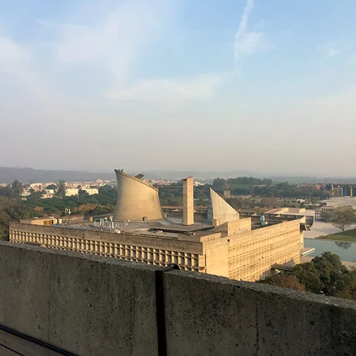 View of the Chandigarh Legislative Assembly building showing its monumental concrete structure, curved roof forms, and surrounding cityscape.