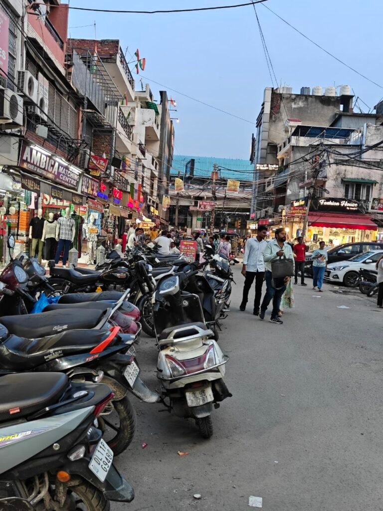 Busy Indian city street lined with small shops, illegally parked two-wheelers and pedestrians walking on the carriageway, highlighting lack of footpaths and parking infrastructure in urban India.