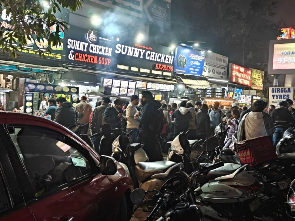 Crowd and parked two-wheelers outside Sunny Chicken food outlets on Jail Road in West Delhi at night