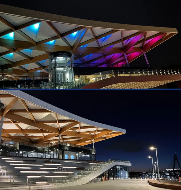 Night view of the Sydney Fish Market’s timber canopy roof illuminated in blue and pink lighting, highlighting its triangular structural geometry above the waterfront.
