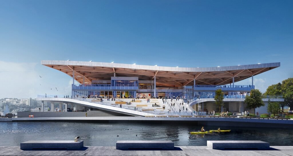 Waterfront view of the Sydney Fish Market featuring stepped public seating, glazed retail levels, and the floating timber roof overlooking Blackwattle Bay.